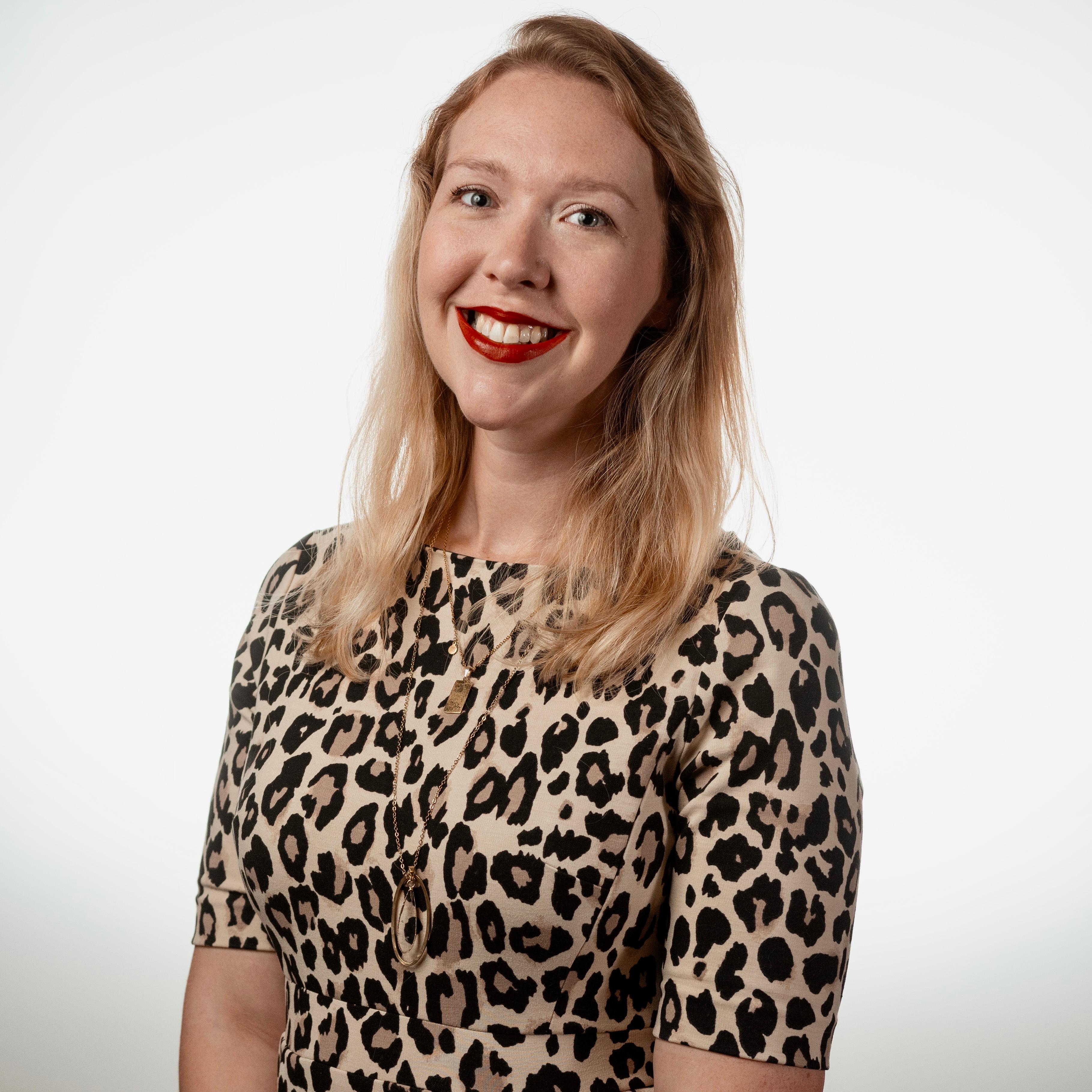 A professional headshot of our speaker Britt Sikora Drake, a young woman with blonde hair wearing a cheetah print long-sleeved shirt and red lipstick in front of a grey brackground.